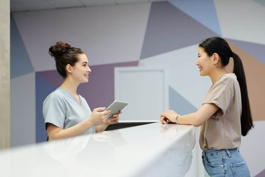A smiling receptionist with a tablet talks with a visitor at a clinic front desk