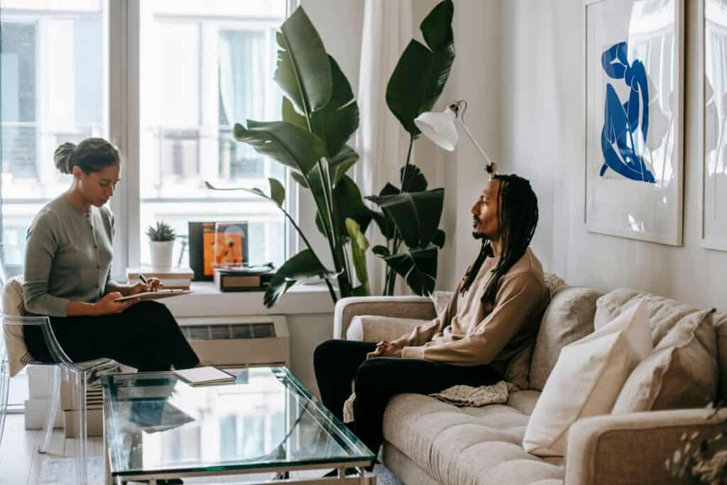 A therapist takes notes while talking with a client on a sofa in a bright room
